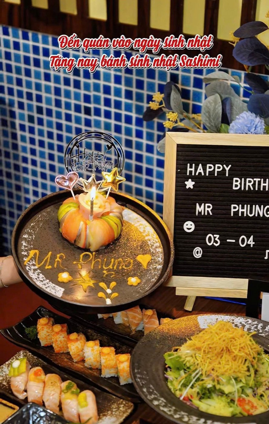 Bánh sinh nhật Sashimi cắm nến và thẻ Happy Birthday trên đĩa ghi tên khách, bên cạnh bảng chúc mừng sinh nhật, khay sushi và salad